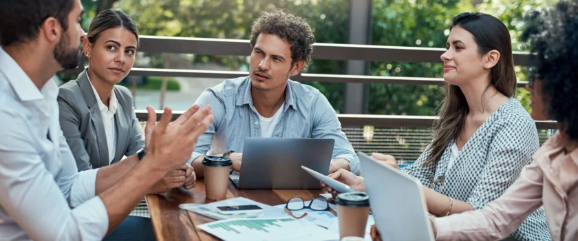 Five people talking around a table with laptops and paper spread out in front of them